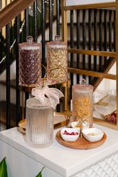 Stylish home countertop display of cut-glass canisters filled with dried herbs and rosebuds on a gold tiered tray and wooden board, set beside a black staircase banister.