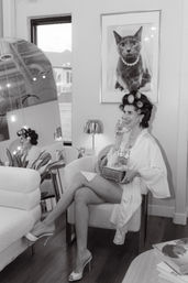 Black-and-white image of a woman in hair rollers and a satin robe sitting in a chic boutique salon chair, smiling while holding a vintage rotary phone; crossed legs in high heels, pearl necklace, framed cat portrait on the wall, mirror reflection and cozy salon decor.