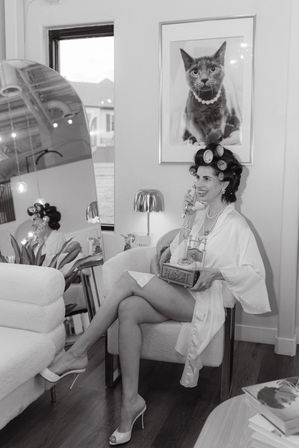 Black-and-white image of a woman in hair rollers and a satin robe sitting in a chic boutique salon chair, smiling while holding a vintage rotary phone; crossed legs in high heels, pearl necklace, framed cat portrait on the wall, mirror reflection and cozy salon decor.