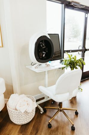 Bright modern spa corner with a circular skin-analysis device on a white desk, computer monitor, plush white rolling chair, woven basket of towels and a potted plant by a glass door.