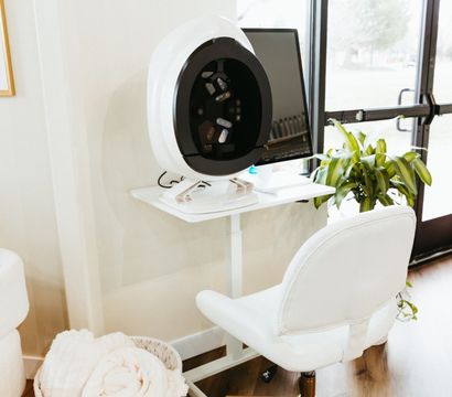 Bright modern spa corner with a circular skin-analysis device on a white desk, computer monitor, plush white rolling chair, woven basket of towels and a potted plant by a glass door.