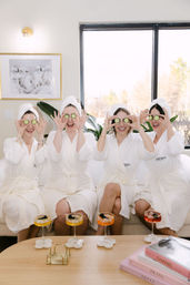 Four friends in white spa robes and towel turbans sit on a bright modern sofa, laughing as they hold cucumber slices over their eyes during a playful at-home spa day with colorful cocktails on the coffee table.