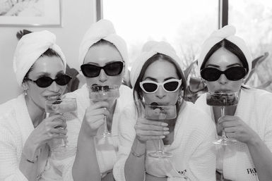 Four friends in white robes and towel turbans wearing sunglasses, sipping berry-garnished champagne in coupe glasses during a playful indoor spa day.