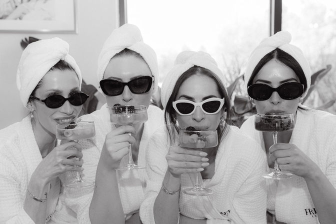 Four friends in white robes and towel turbans wearing sunglasses, sipping berry-garnished champagne in coupe glasses during a playful indoor spa day.