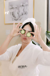 Person in a white bathrobe with a towel wrapped around their hair, playfully holding cucumber slices over their eyes and pouting in a spa-like bathroom setting with framed art and plants.