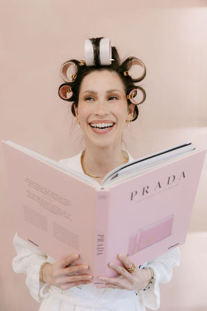 Laughing woman with pink hair rollers and gold earrings reading a pastel pink fashion coffee-table book against a soft pink backdrop — playful beauty-at-home moment