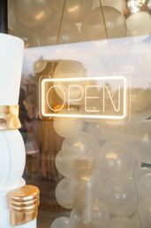 Glowing neon "OPEN" sign hanging in a storefront window, layered over cream balloons and gold-accented decor in a festive shop display.