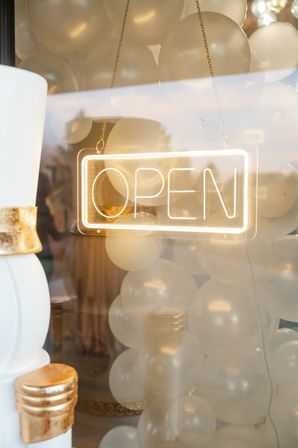 Glowing neon "OPEN" sign hanging in a storefront window, layered over cream balloons and gold-accented decor in a festive shop display.