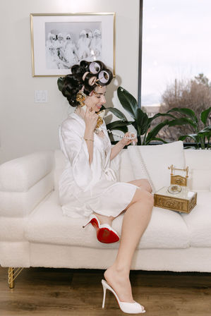 Retro-glam getting-ready scene: woman in a white satin robe with hair rollers sitting on a plush white sofa, smiling while talking on a gold vintage rotary phone, wearing white high-heeled mules with red soles, indoor living room with plants and framed art.