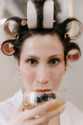 Close-up of woman with pink hair rollers and clips sipping a golden drink with blackberries from a gold-rimmed glass