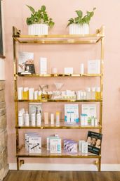 Chic gold shelving on a blush pink wall displaying potted plants and neatly arranged skincare and beauty products in a boutique spa or salon retail display.