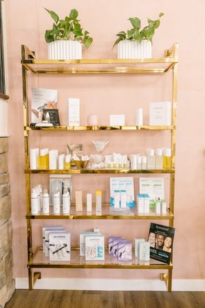 Chic gold shelving on a blush pink wall displaying potted plants and neatly arranged skincare and beauty products in a boutique spa or salon retail display.