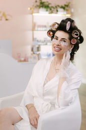 Smiling woman in a boutique beauty salon chair with pink hair rollers and under-eye patches, wearing a white robe, pearl necklace and lace glove during a pampering hair and facial treatment.