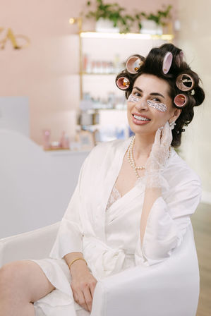 Smiling woman in a boutique beauty salon chair with pink hair rollers and under-eye patches, wearing a white robe, pearl necklace and lace glove during a pampering hair and facial treatment.