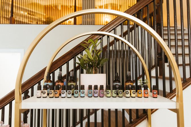 Colorful essential oil bottles and spray misters lined on a white shelf with gold arched frame and a potted plant, displayed in front of a wooden staircase in a modern home interior.