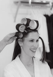 Black-and-white portrait of a smiling woman in a salon with large hair rollers being clipped in by a stylist during a styling session.