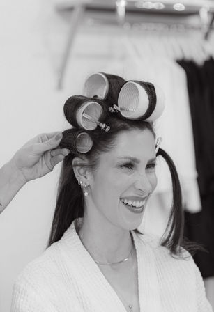 Black-and-white portrait of a smiling woman in a salon with large hair rollers being clipped in by a stylist during a styling session.