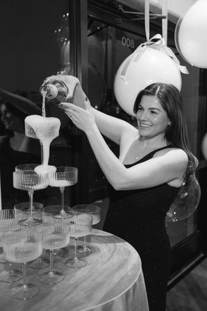 Smiling woman in a black dress pouring champagne into a cascading coupe tower at an indoor celebration with large ribboned balloons