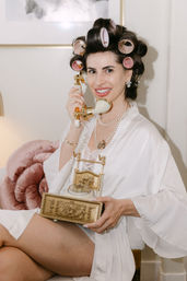 Smiling woman in a white satin robe with pink hair rollers holding an ornate gold rotary phone in a cozy boudoir setting