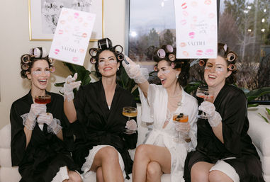 Four laughing women in white and black robes with hair rollers, lace gloves, and under-eye patches sit on a sofa toasting with cocktails and toss a lipstick-kiss print card during a playful at-home pampering party.