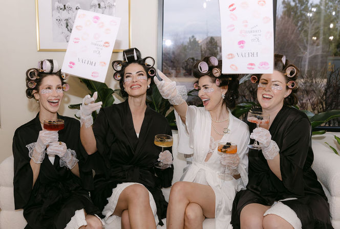 Four laughing women in white and black robes with hair rollers, lace gloves, and under-eye patches sit on a sofa toasting with cocktails and toss a lipstick-kiss print card during a playful at-home pampering party.
