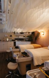 Relaxing modern spa pedicure room with two pedicure stations and plush treatment beds beneath a flowing white draped ceiling, warm ambient lighting, wooden floors, shelves of skincare products and potted palms.