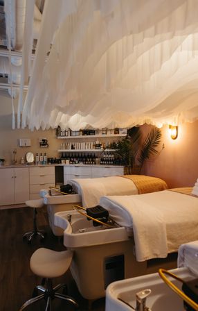 Relaxing modern spa pedicure room with two pedicure stations and plush treatment beds beneath a flowing white draped ceiling, warm ambient lighting, wooden floors, shelves of skincare products and potted palms.