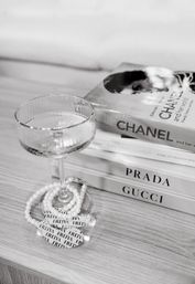 Monochrome coffee-table vignette featuring a textured coupe glass, pearl necklace, and stacked designer fashion books on a light wood surface.