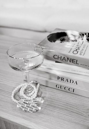 Monochrome coffee-table vignette featuring a textured coupe glass, pearl necklace, and stacked designer fashion books on a light wood surface.