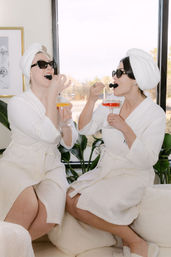 Two friends on a relaxing spa day in white robes and towel turbans, wearing sunglasses and sharing blackberries and colorful cocktails in a bright indoor spa lounge by a large window