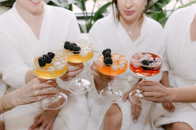 Four friends in white spa robes toasting with colorful, blackberry‑garnished cocktails in coupe glasses — bright spa‑day celebration