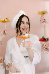 Smiling woman in a white spa robe and towel turban toasting with champagne coupes held by friends against a blush pink backdrop — spa day celebration.