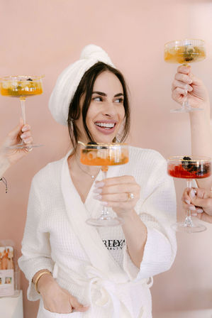 Smiling woman in a white spa robe and towel turban toasting with champagne coupes held by friends against a blush pink backdrop — spa day celebration.