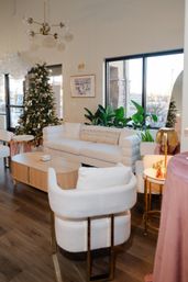 Cozy modern living room with white boucle sofa and chairs, light wood coffee table, decorated Christmas tree with white and gold ornaments, large windows, indoor plants and brass accents.