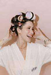 Smiling woman in a white spa robe wearing pink and purple hair rollers and gold jewelry, adjusting curl clips against a blush-pink background — playful beauty routine.