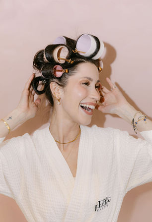 Smiling woman in a white spa robe wearing pink and purple hair rollers and gold jewelry, adjusting curl clips against a blush-pink background — playful beauty routine.