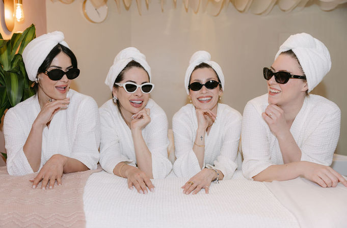 Four women in white robes with towel turbans and sunglasses, smiling and chatting while leaning on a spa treatment table during a pampering day.