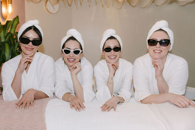 Four women in white spa robes and towel turbans wearing sunglasses, smiling and leaning on a treatment table in a bright indoor spa — playful spa day group.