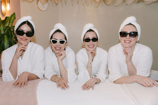 Four women in white spa robes and towel turbans wearing sunglasses, smiling and leaning on a treatment table in a bright indoor spa — playful spa day group.