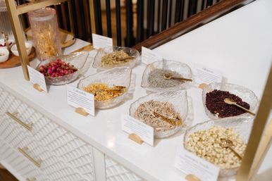 Leaf-shaped glass bowls holding dried herbs and flower teas (rosebuds, calendula, lavender, hibiscus) with gold scoops on a white countertop — boutique herbal tea display.