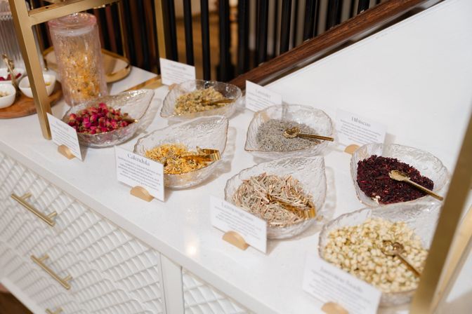 Leaf-shaped glass bowls holding dried herbs and flower teas (rosebuds, calendula, lavender, hibiscus) with gold scoops on a white countertop — boutique herbal tea display.