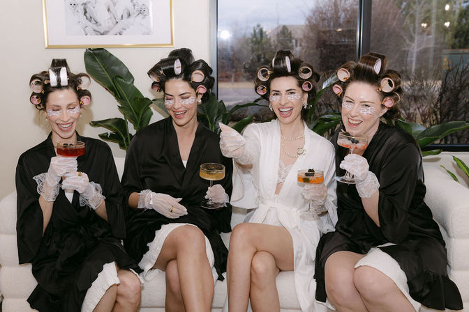 Four women in robes with hair rollers, under-eye patches and lace gloves laughing on a couch while holding cocktails during a playful pampering/getting-ready party in a bright lounge with plants and window view.