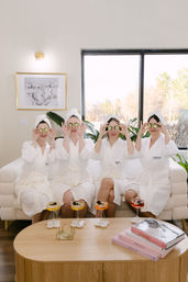Four women in white robes and towel turbans holding cucumber slices over their eyes during a light-filled indoor spa day on a plush white sofa, with cocktails and stacked style books on a wooden coffee table and large window views.