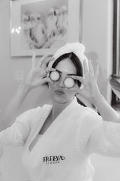 Playful woman in a white spa robe and towel turban holding cucumber slices over her eyes during an at-home skincare moment
