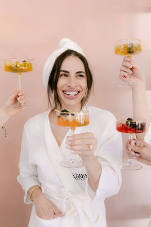 Spa day celebration: smiling woman in a white robe and towel turban toasting colorful champagne coupe cocktails with friends against a soft pink backdrop