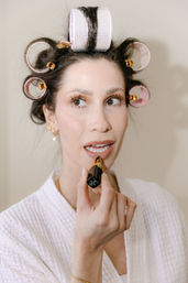 Close-up of woman in white robe with pink hair rollers applying nude lipstick during at-home beauty routine