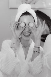Playful woman in a white robe and towel turban holding cucumber slices over her eyes during a spa facial and skincare routine