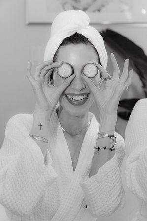 Playful woman in a white robe and towel turban holding cucumber slices over her eyes during a spa facial and skincare routine