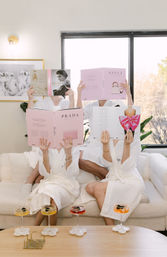 Three friends in white spa robes lounging on a cream sofa in a bright living room, each holding pink fashion books over their faces, with colorful cocktails on a wooden coffee table and a large window behind them.