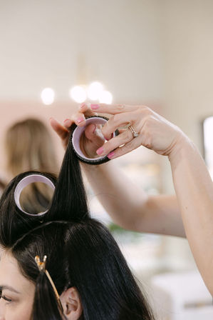 Close-up of hair styling at a beauty salon: stylist’s hands with pink nails and a ring rolling long dark hair into lavender velcro rollers, hair section clipped and salon lights blurred in background.